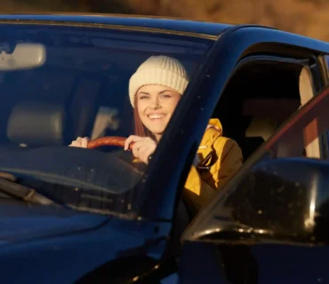 Mujer joven sonriendo mientras conduce un coche negro en un entorno natural al atardecer.