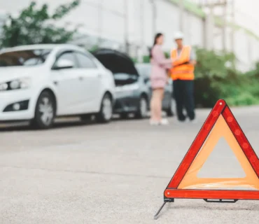 Señal de emergencia triangular colocada en el suelo con coches averiados y una mujer hablando con un operario al fondo.