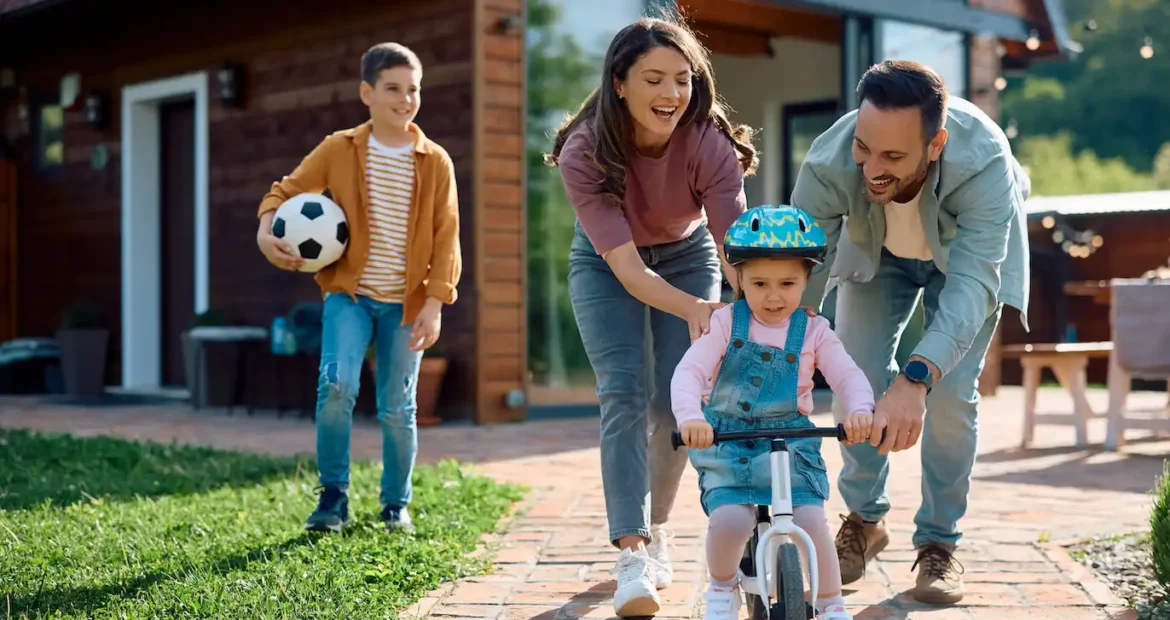 Familia jugando en el jardín frente a su casa mientras los padres ayudan a su hija pequeña a montar en bicicleta, concepto de protección y seguridad en el hogar.
