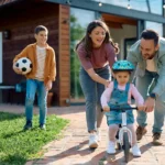 Familia jugando en el jardín frente a su casa mientras los padres ayudan a su hija pequeña a montar en bicicleta, concepto de protección y seguridad en el hogar.