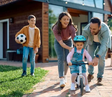 Familia jugando en el jardín frente a su casa mientras los padres ayudan a su hija pequeña a montar en bicicleta, concepto de protección y seguridad en el hogar.