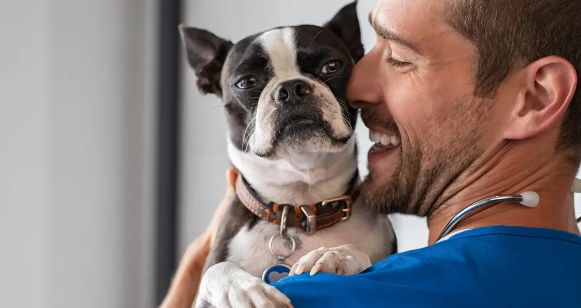 Un hombre alegre, con el pelo castaño corto y barba, sonríe mientras sostiene en brazos a un perro blanco y negro.