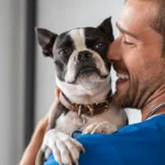 Un hombre alegre, con el pelo castaño corto y barba, sonríe mientras sostiene en brazos a un perro blanco y negro.